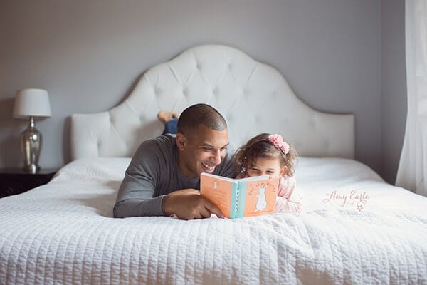 Dad reading to daughter while lying on a queen platform bed with upholstered headboard. There is a nightstand to the side of the bed.